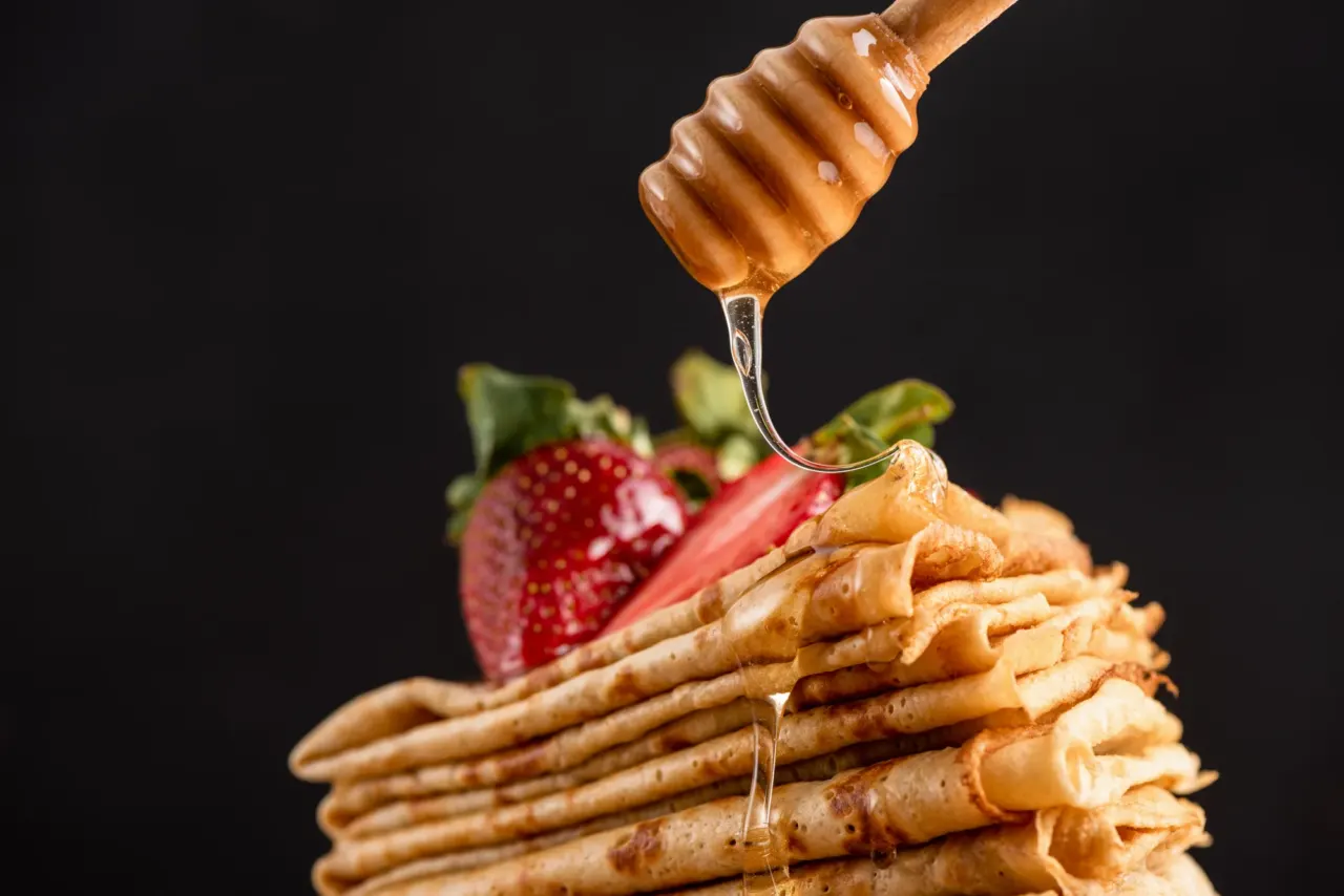 Pile de crêpes aux fraises et miel coulant d'une cuillère en bois