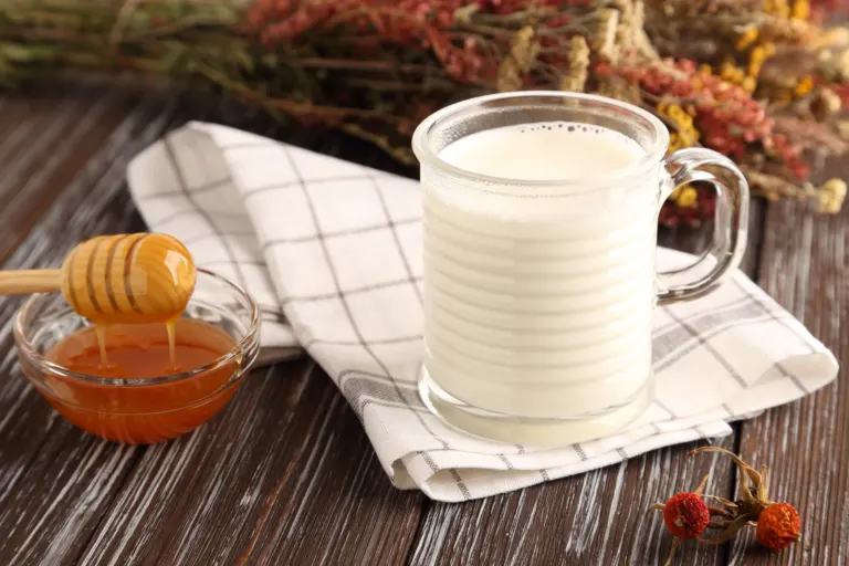 Grande tasse en verre de lait chaud sur un torchon à carreaux, petit bol de miel et cuillère en bois