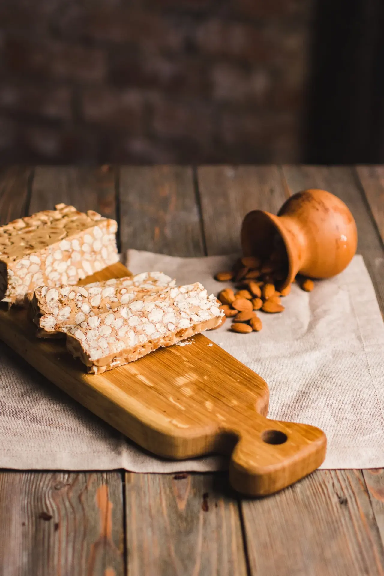 Tranches de nougat blanc aux amandes sur une planche en bois, amandes débordant d'un petit pot en terre cuite