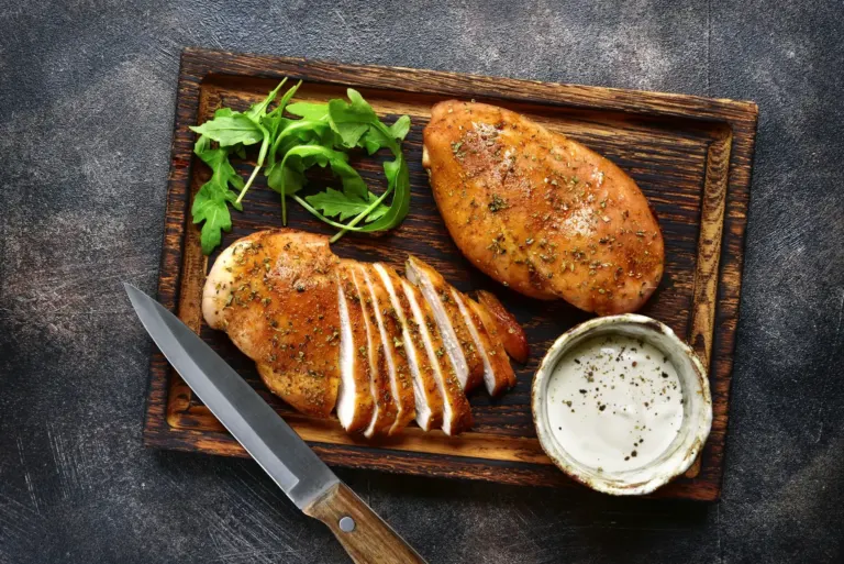 Two golden chicken fillets on a wooden board, one sliced, with rocket and a ramekin of creamy sauce