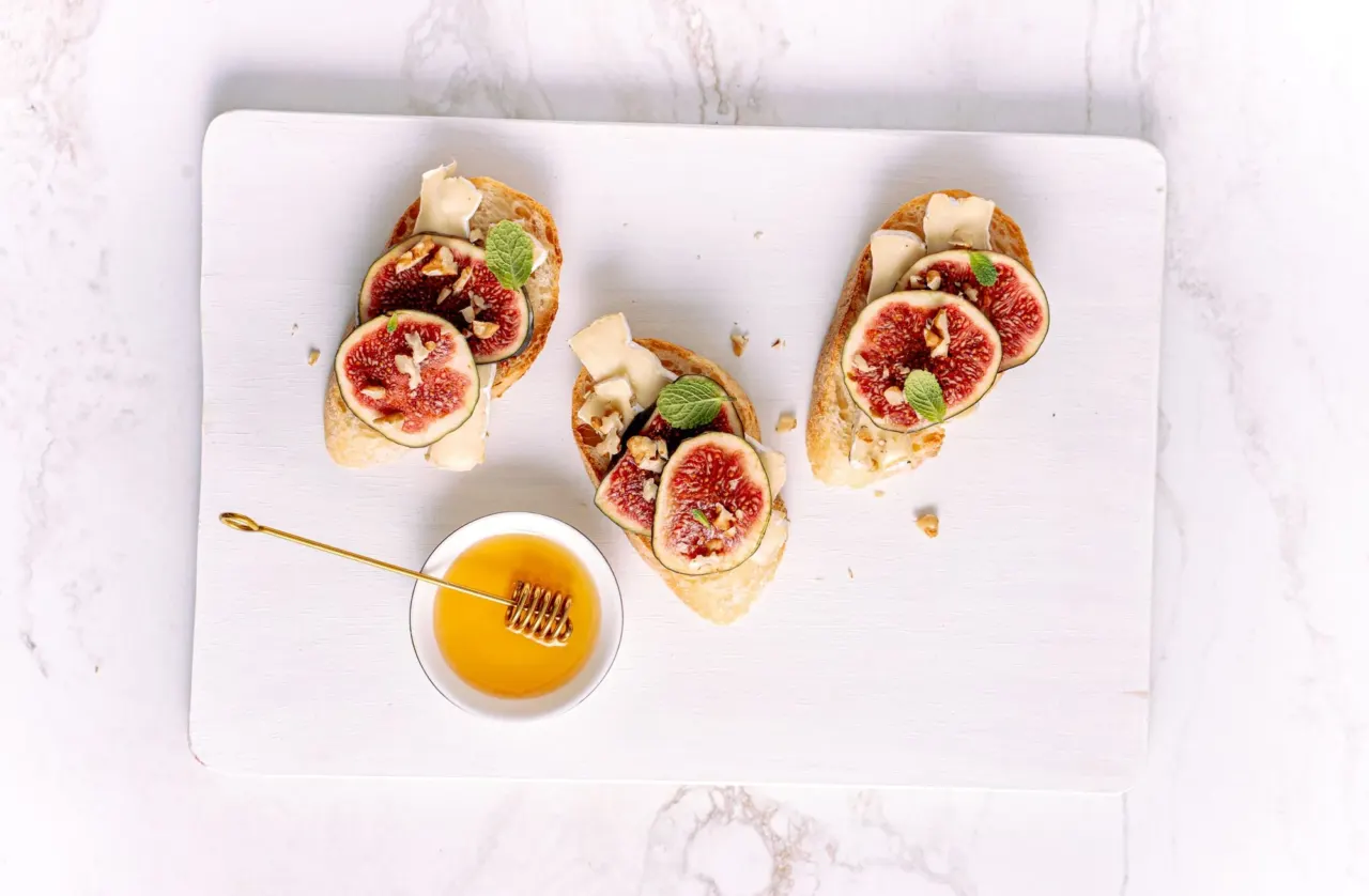Trois toasts de chèvre frais et figues coupées en deux, près d'un bol de miel et d'une cuillère à miel